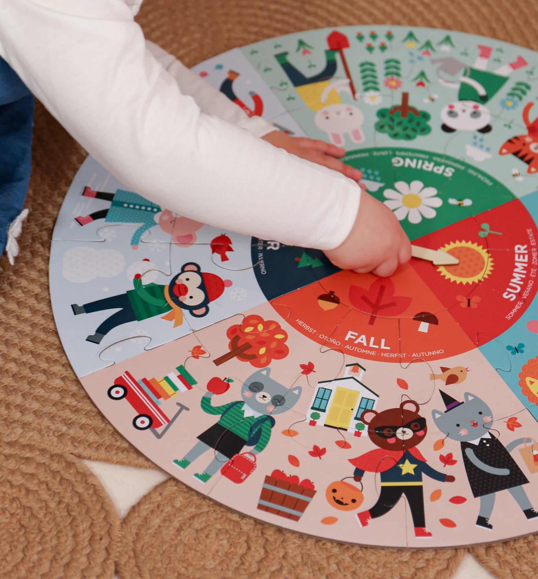 A child’s hand points to the Fall section of the Petit Collage Seasons 25-Piece Floor Puzzle, a colorful circular chart featuring animals and seasonal activities.