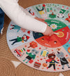 A child’s hand points to the Fall section of the Petit Collage Seasons 25-Piece Floor Puzzle, a colorful circular chart featuring animals and seasonal activities.