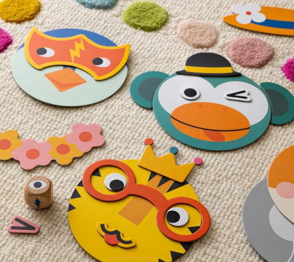 Petit Collage’s Make Funny Faces Game features colorful animal face puzzles and accessories, like a tiger with glasses and a crown, arranged on a beige textured carpet with multicolored dots.