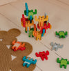 The Petit Collage Animal Acrobats Balance Game features colorful wooden animal figures stacked on a white floor, with some scattered nearby and a textured rug partially visible.
