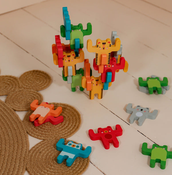 The Petit Collage Animal Acrobats Balance Game features colorful wooden animal figures stacked on a white floor, with some scattered nearby and a textured rug partially visible.