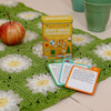 A tin labeled Busy Ideas for Bored Kids Joyful Edition by Petit Collage sits on a green crocheted blanket, with an apple, two green cups, and activity cards nearby.