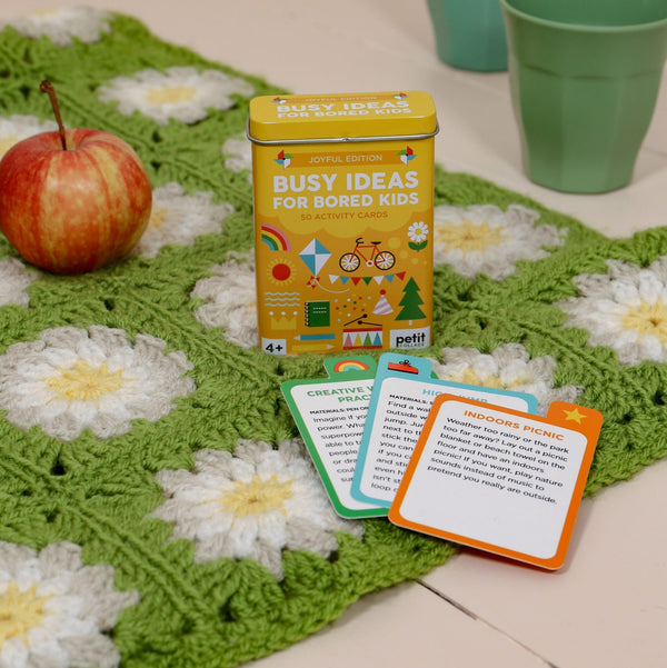 A tin labeled Busy Ideas for Bored Kids Joyful Edition by Petit Collage sits on a green crocheted blanket, with an apple, two green cups, and activity cards nearby.