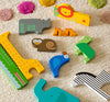 The Petit Collage Wooden Puzzle + Play Shaped Safari Animals features a giraffe, crocodile, rhino, lion, monkey, bird, zebra, turtle, and tiger playfully arranged on a colorful polka dot textured carpet.