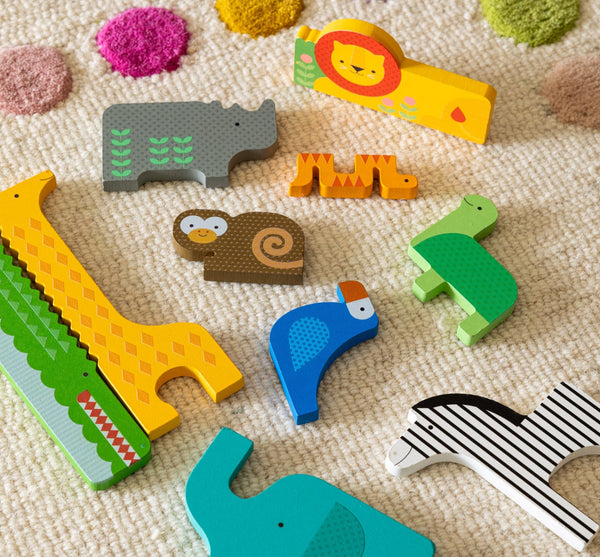 The Petit Collage Wooden Puzzle + Play Shaped Safari Animals features a giraffe, crocodile, rhino, lion, monkey, bird, zebra, turtle, and tiger playfully arranged on a colorful polka dot textured carpet.