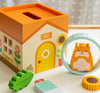 The Petit Collage Petit Pals: Hugo Hamster Wooden Toy features a wooden house with a yellow door, colorful toy pieces like a green leaf and sunflower, and Hugo the hamster in a blue ring.