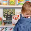 A young child in a blue dress holds the Petit Collage Shake and Play: Wooden Tambourine, standing in front of a bookshelf filled with childrens books and toys.