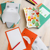 The Festive Pals My Stationery Set by Petit Collage features animal-themed notecards, envelopes, lined paper, stickers with trees and animals, plus colored pencils—all displayed on a wooden surface.