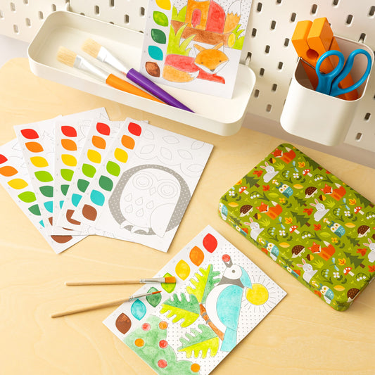 The desk displays the Petit Collage Paint by Numbers: Woodland kit, with paint brushes, watercolor palettes, scissors, a forest-patterned pencil case, and finished nature-inspired artwork.
