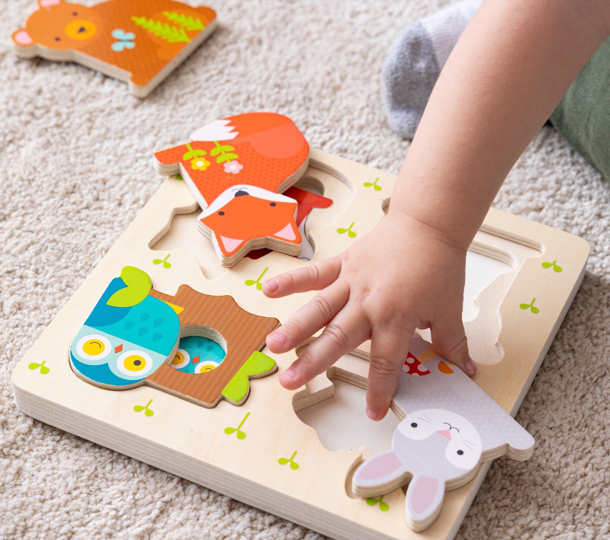 A childs hand places a wooden piece into the Reveal Puzzle: You Are My Baby by Petit Collage, matching it to an illustrated animal slot on the board.