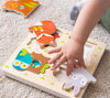 A childs hand places a wooden piece into the Reveal Puzzle: You Are My Baby by Petit Collage, matching it to an illustrated animal slot on the board.