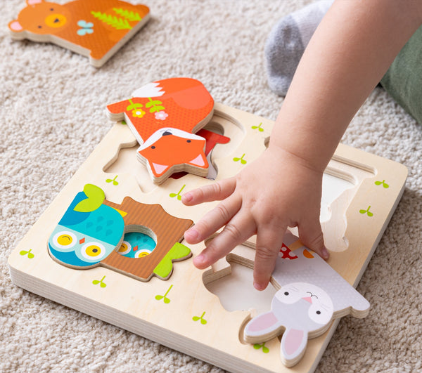 A childs hand places a wooden piece into the Reveal Puzzle: You Are My Baby by Petit Collage, matching it to an illustrated animal slot on the board.