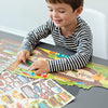 A young child in a striped shirt plays at a table with the Petit Collage Roads & Rails Sticker Activity Set, which includes a colorful city-themed mat and stickers.