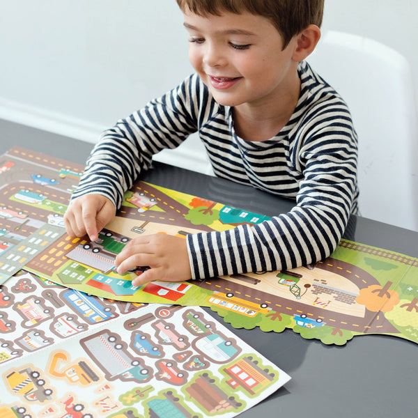 A young child in a striped shirt plays at a table with the Petit Collage Roads & Rails Sticker Activity Set, which includes a colorful city-themed mat and stickers.