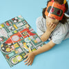 A child in a striped shirt and robot mask sits on a blue floor beside the completed Petit Collage Robot Factory 100-Piece Decoder Puzzle, resting one hand on the puzzle.