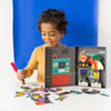 A young child arranges pieces from the Petit Collage Superheroes Magnetic Dress Up Play Set on a table, with the open box showing two superhero figures.