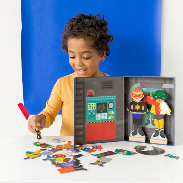 A young child arranges pieces from the Petit Collage Superheroes Magnetic Dress Up Play Set on a table, with the open box showing two superhero figures.