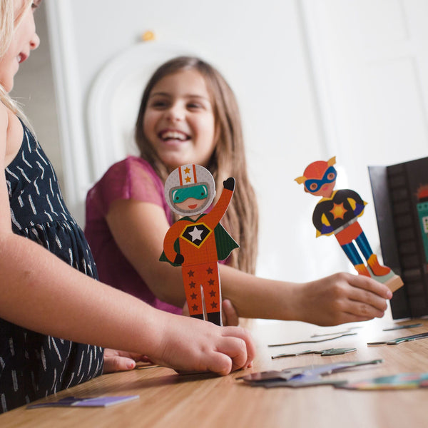 Two children smile and play at a table with the Petit Collage Superheroes Magnetic Dress Up Play Set.