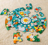 The Petit Collage Telling the Time 25-Piece Floor Puzzle, a colorful animal-themed circular puzzle with a numbered clock at its center, is shown incomplete with several pieces scattered on a beige carpet.