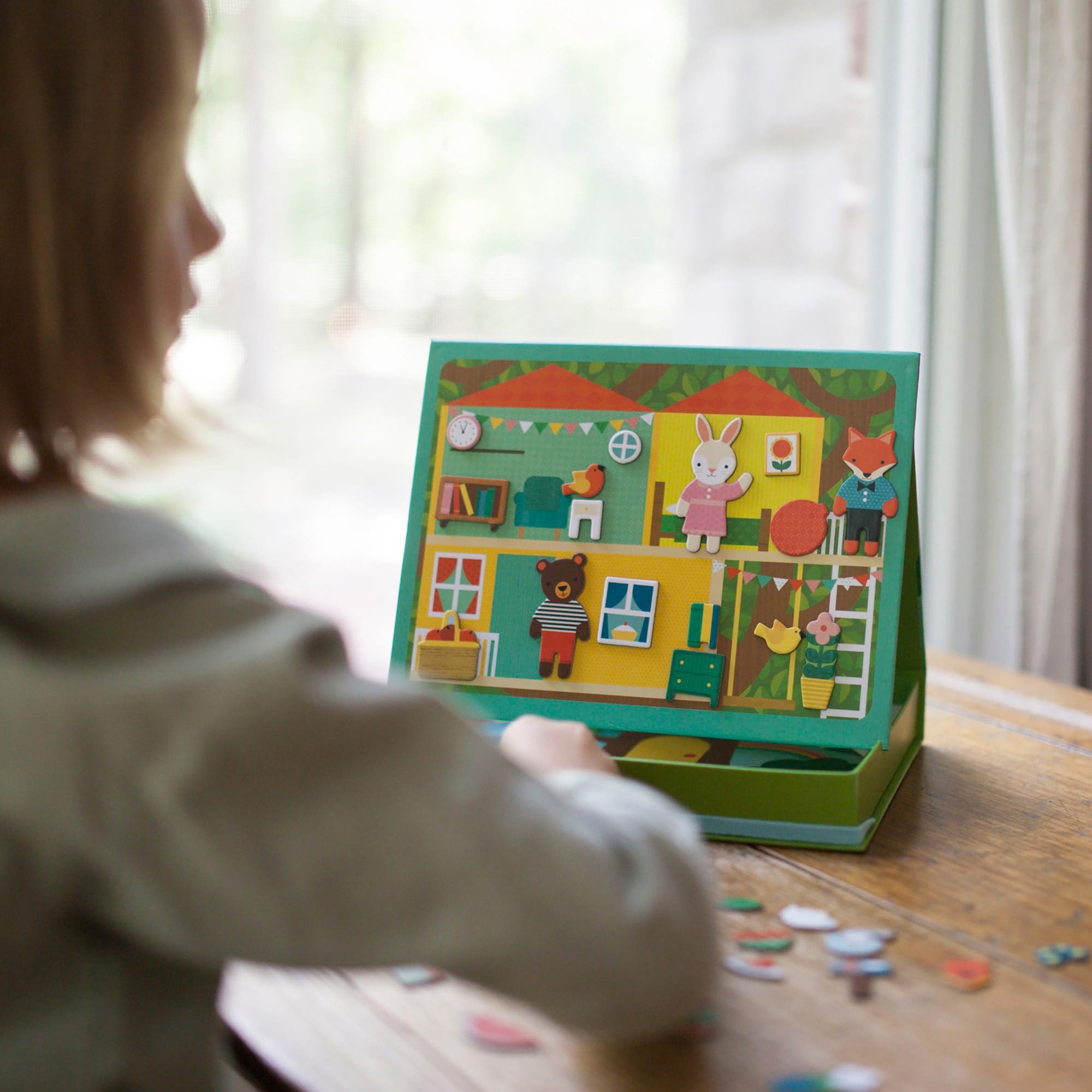 A child sits at a table playing with the Petit Collage Treehouse Party Magnetic Play Scene, arranging magnetic pieces on the illustrated background.