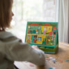 A child sits at a table playing with the Petit Collage Treehouse Party Magnetic Play Scene, arranging magnetic pieces on the illustrated background.