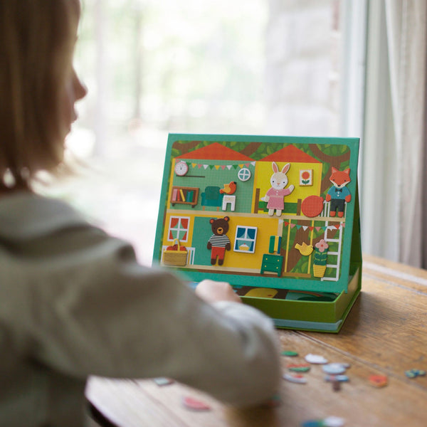A child sits at a table playing with the Petit Collage Treehouse Party Magnetic Play Scene, arranging magnetic pieces on the illustrated background.