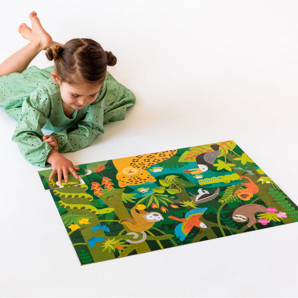 A young girl in a green dress assembles the Petit Collage Wild Rainforest 24-Piece Floor Puzzle, which features colorful jungle animals like a jaguar, sloth, and toucan.