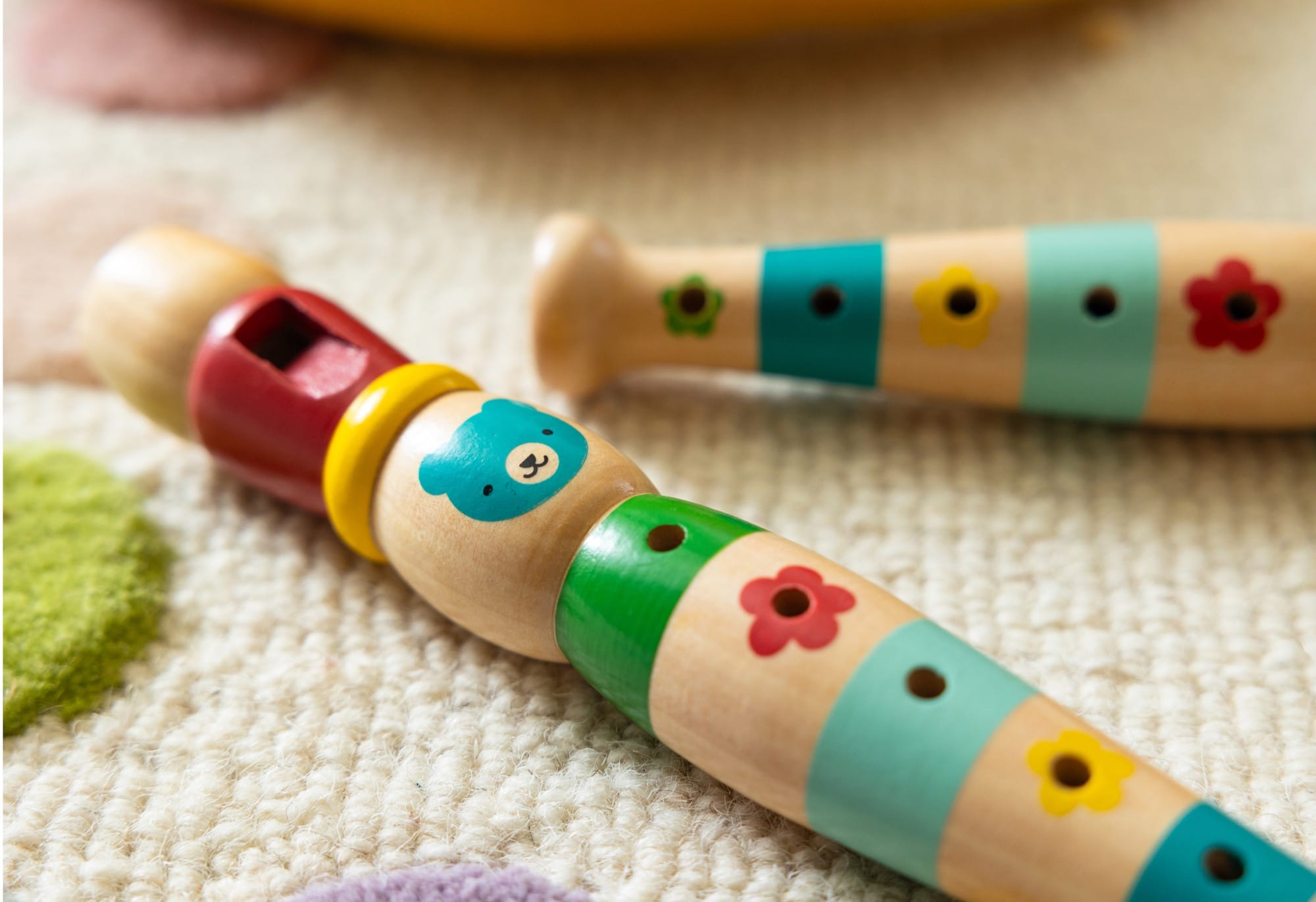 Two Petit Collage Playful Tunes Wooden Recorders with vibrant bear and flower designs are placed on a textured carpet.