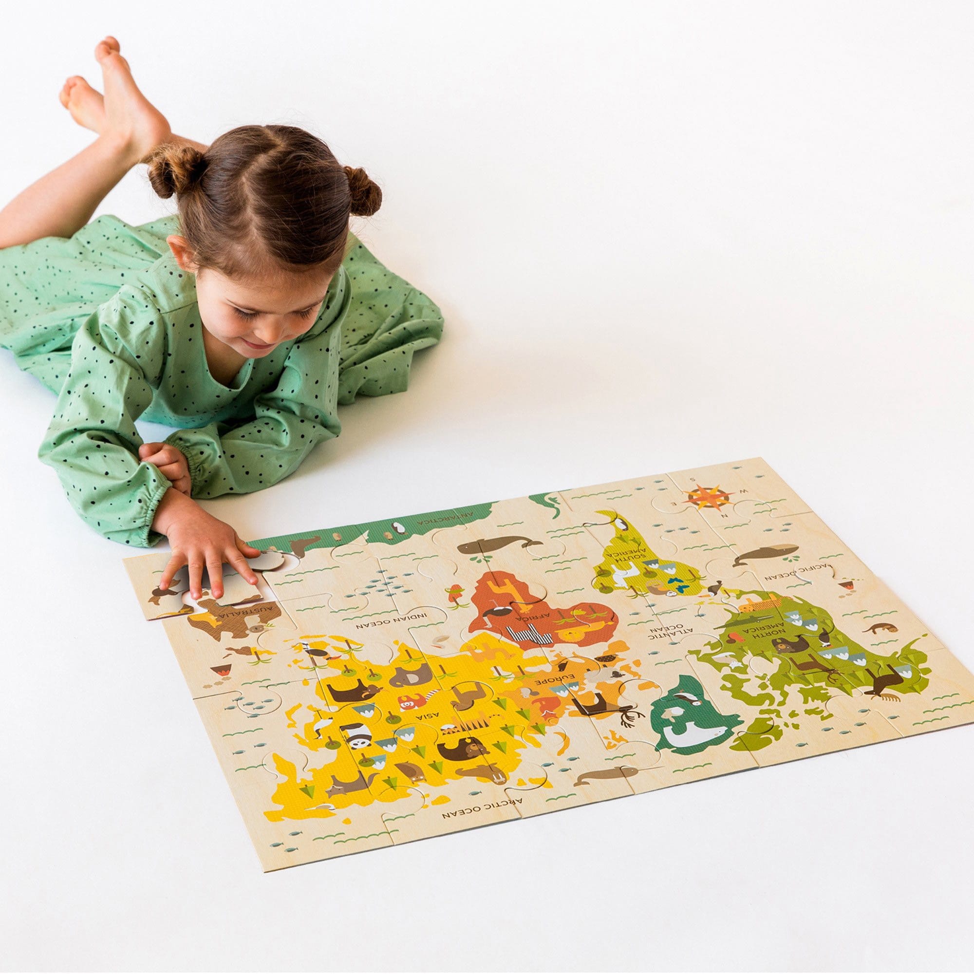 A young child in a green dress lies on the floor, assembling the vibrant World Map 24-Piece Floor Puzzle by Petit Collage.