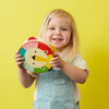 A young child with blonde hair holds the Petit Collage Kaleidoscope Wooden Lion Tambourine, a colorful toy featuring a lion face, and stands in front of a yellow background.