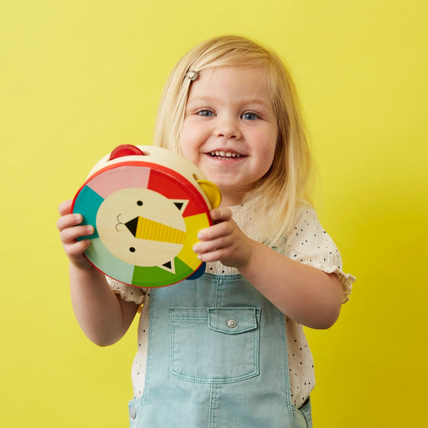A young child with blonde hair holds the Petit Collage Kaleidoscope Wooden Lion Tambourine, a colorful toy featuring a lion face, and stands in front of a yellow background.
