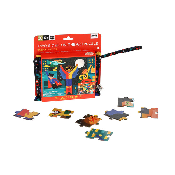 The Petit Collage Two Sided Superheroes On-the-Go Puzzle for ages 5+ is shown partly assembled with loose pieces, in front of its colorful, portable packaging featuring a handle.