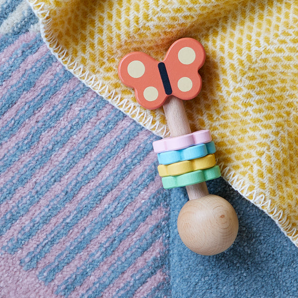The Petit Collage Butterfly Wooden Shaker, featuring a butterfly design and colorful rings, sits on a blue and pink textured rug beside a yellow patterned blanket.