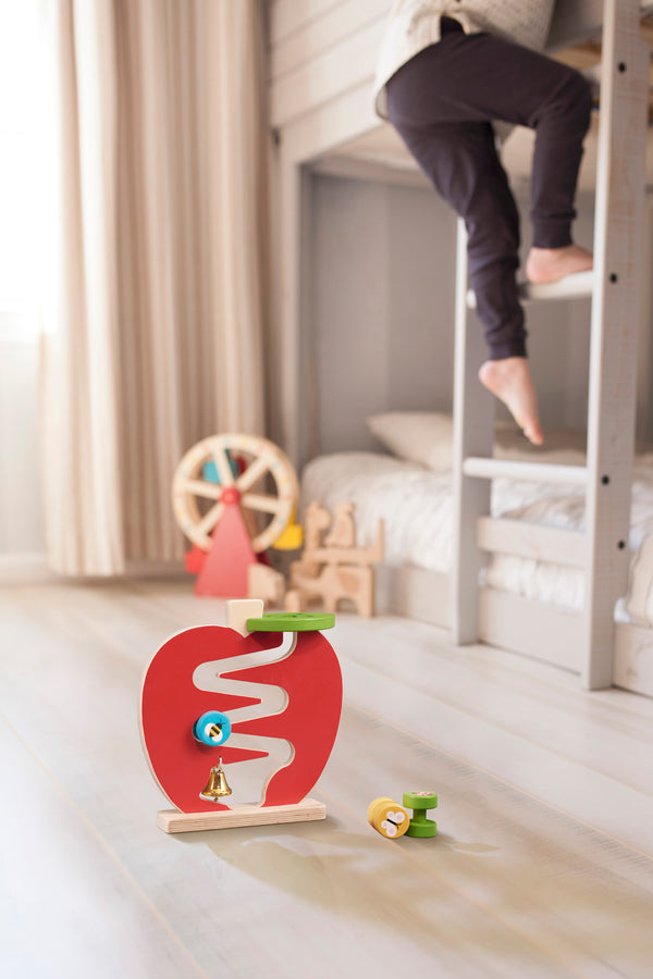 A child climbs down a bunk bed ladder in a bright room, where the Petit Collage Wooden Apple Run Play Set and other toys are scattered on the floor.