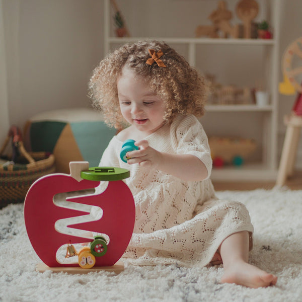 A young child with curly hair plays with the Petit Collage Wooden Apple Run Play Set on a rug in a living room.