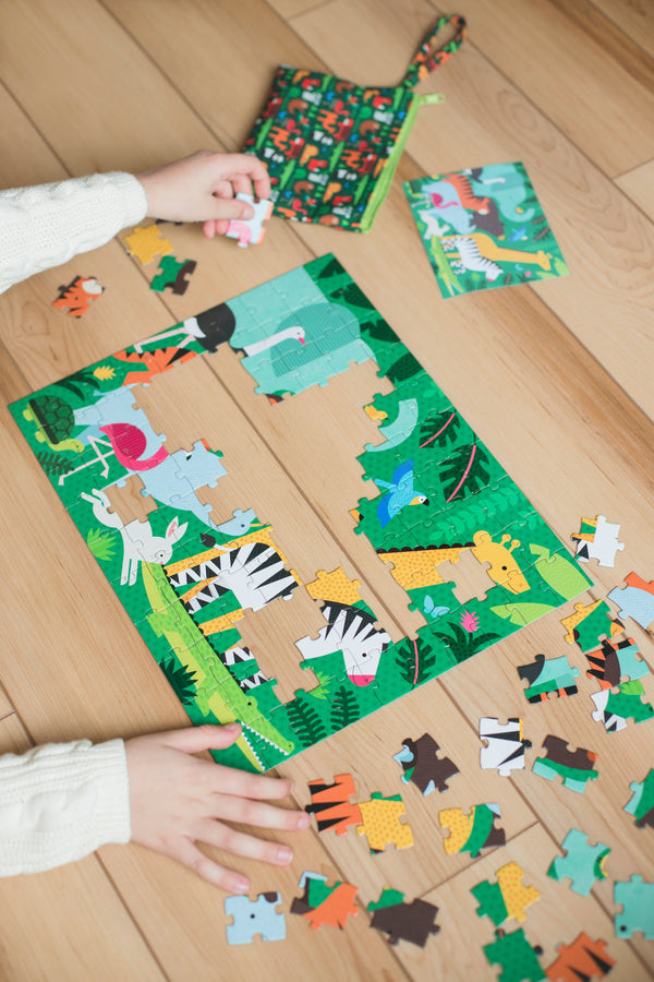A child assembles the Petit Collage Two Sided Animal Menagerie On-The-Go Puzzle on a wooden floor, with colorful animal pieces and the included small storage bag nearby.