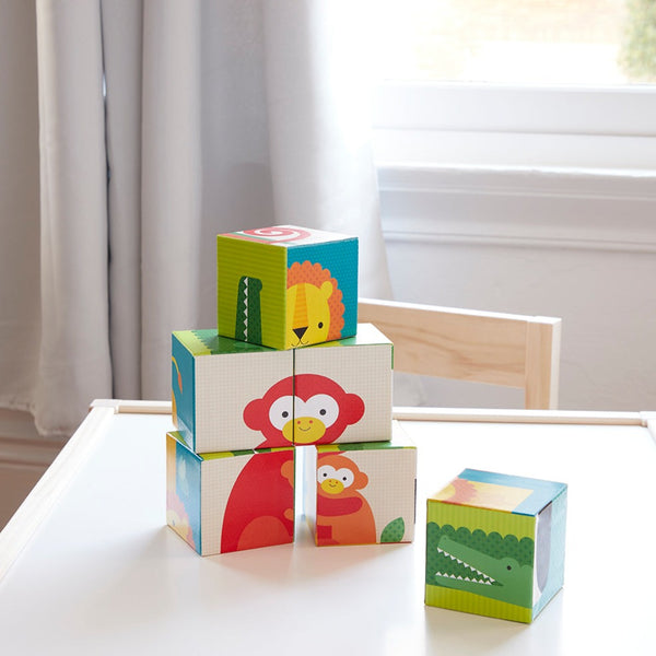 Four vibrant Puzzle Blocks - Wild Animals by Petit Collage, featuring animal illustrations, are stacked on a white table near a sunlit window with sheer curtains.