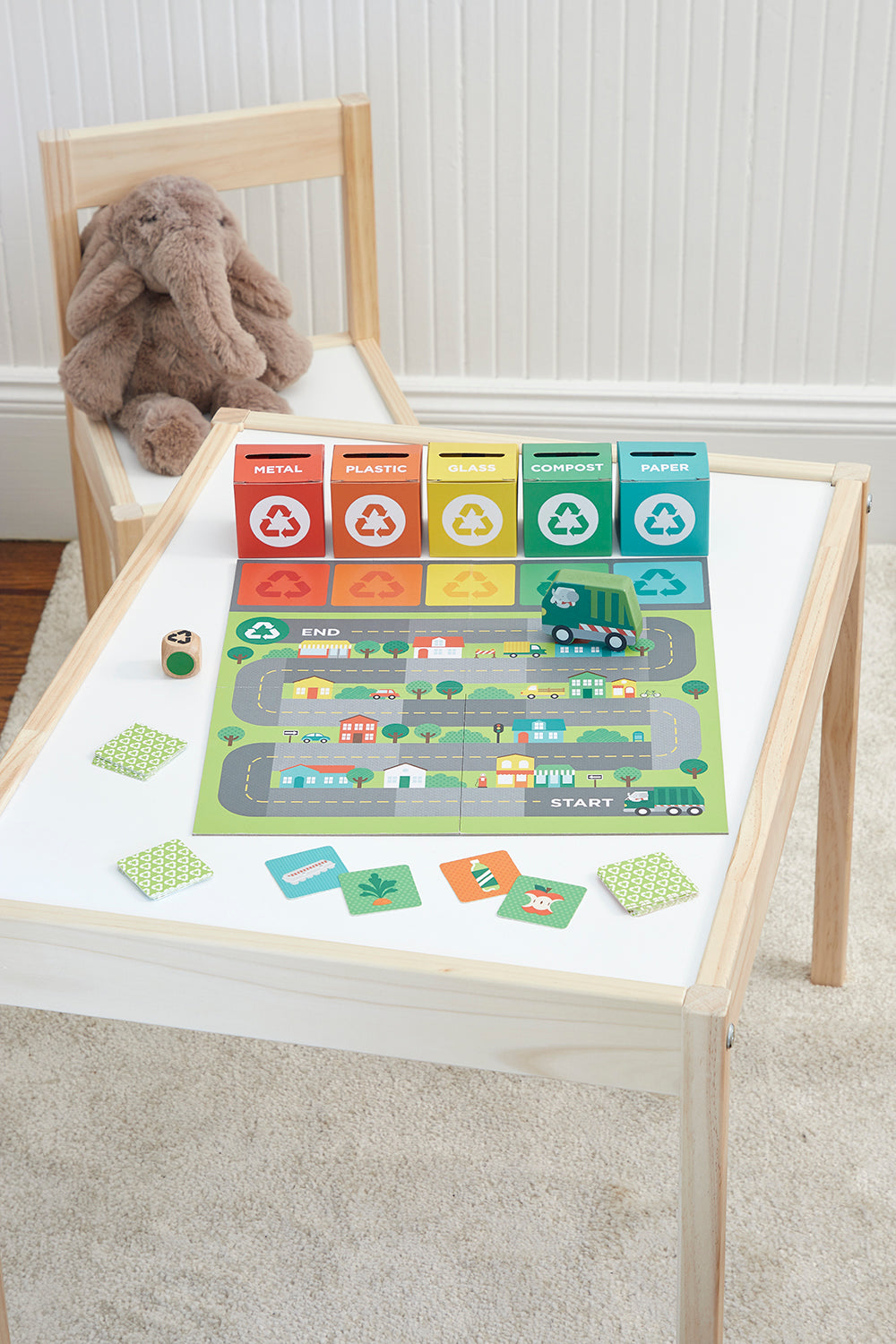 The Petit Collage Rush to Recycle childrens board game, featuring colored bins, a toy truck, cards, and dice, is set up on a small white table beside a wooden chair with a plush bunny.