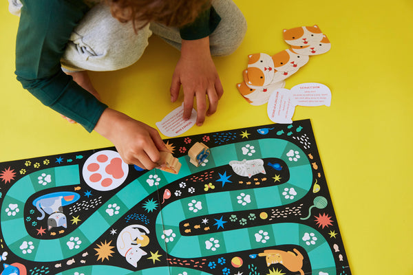 A child plays Petit Collage’s Catventures Board Game on a yellow surface, moving a piece along the winding path with cards and game elements scattered nearby.