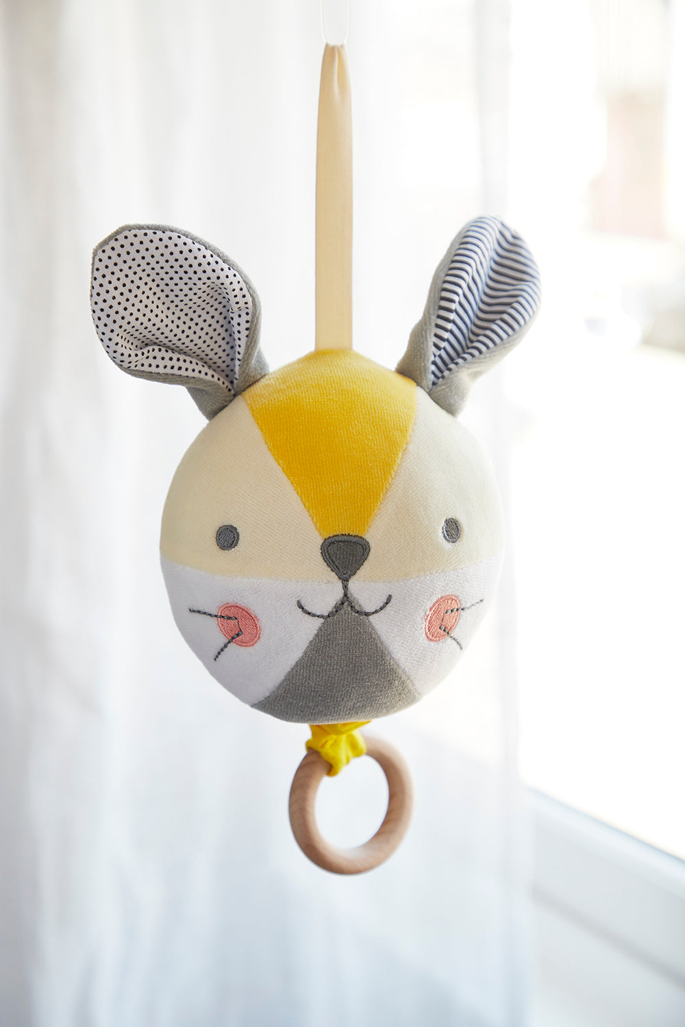 The Petit Collage Busy Bunny Musical Mobile features a soft, round bunny face with large ears, a stitched smile, and a wooden ring, hanging by a fabric loop in front of a bright window.