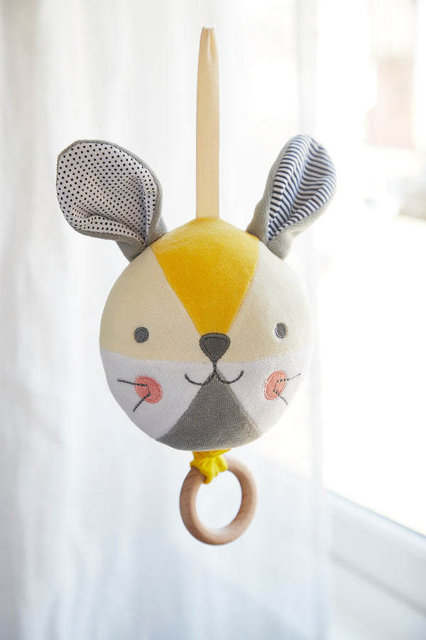 The Petit Collage Busy Bunny Musical Mobile features a soft, round bunny face with large ears, a stitched smile, and a wooden ring, hanging by a fabric loop in front of a bright window.