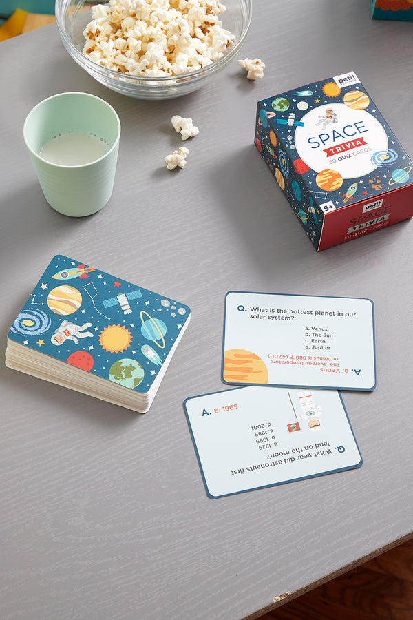A deck of Petit Collage’s Trivia Cards - Space rests on a gray table beside popcorn and milk. Two cards are face up, displaying questions and answers about planets and astronauts.