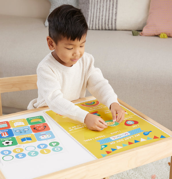 A young boy sits at a wooden table, interacting with the Petit Collage First & Last Day of School Sticker Sign, a colorful educational poster featuring numbers, animals, and shapes.