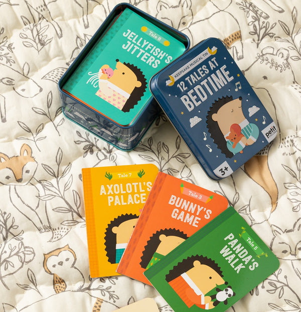 Four illustrated children’s books and two tins, including the Petit Collage “12 Tales at Bedtime,” are displayed on animal-patterned bedding.
