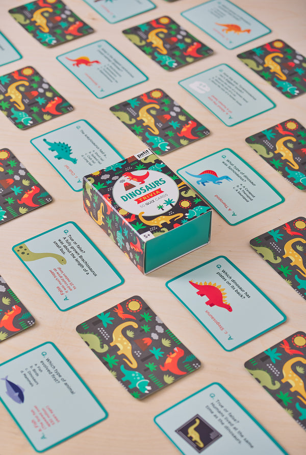 The Dinosaurs Trivia Quiz Cards by Petit Collage are shown with their box surrounded by several dinosaur-themed quiz cards spread out on a wooden surface.