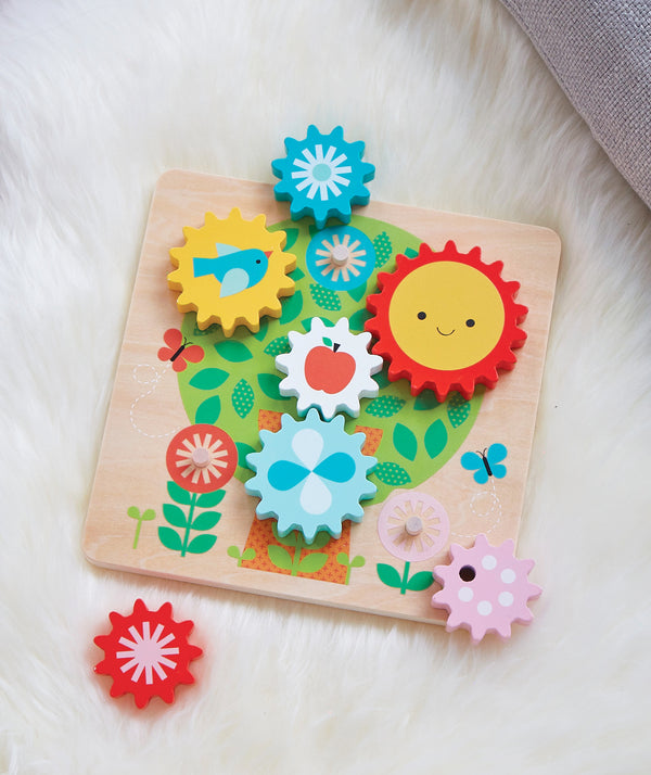 The Petit Collage Busy Tree Wooden Twist Puzzle features colorful, gear-shaped pieces with images like a sun, bird, apple, flower, and abstract patterns on a decorated board displayed on a white furry surface.