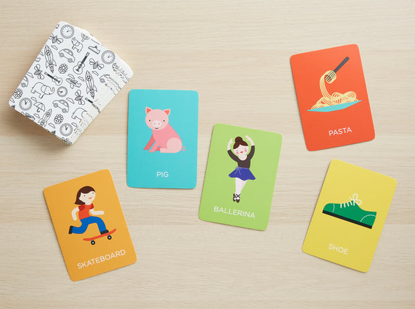 Five illustrated Charades for Kids flashcards by Petit Collage—featuring a pig, ballerina, pasta, skateboard, and shoe—are arranged on a wooden surface with a stack of black and white cards in the corner.