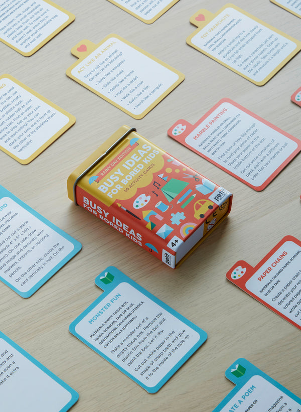 A box labeled Busy Ideas for Bored Kids: Rainy Day Edition by Petit Collage sits on a wooden table, surrounded by colorful activity cards featuring fun prompts and instructions.