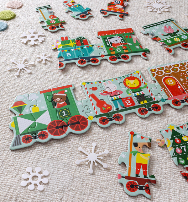 The Petit Collage Christmas Train Shaped Floor Puzzle, featuring vibrant animal designs, is arranged on a textured rug with loose pieces and scattered white paper snowflakes nearby.