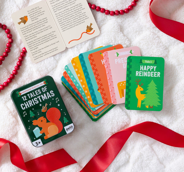 The Petit Collage 12 Tales of Christmas set features colorful animal-illustrated story cards, displayed on a white blanket with red ribbon and beads. An open booklet from the set is also shown.